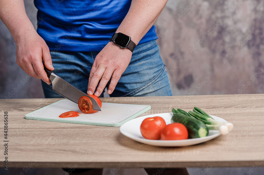 A man cuts a red tomato on a board. Cucumbers and tomatoes in a white plate