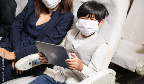 Asian boy playing on digital tablet at seat in airplane cabin. passenger wearing medical face mask for self-prevention Covid-19 during travel abroad. new normal concept