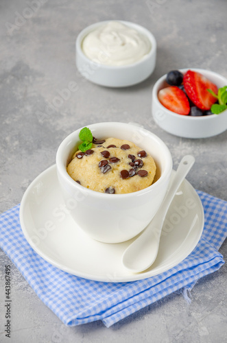Mug cake with chocolate chips, fresh berries and cream on a gray concrete background. Copy space.