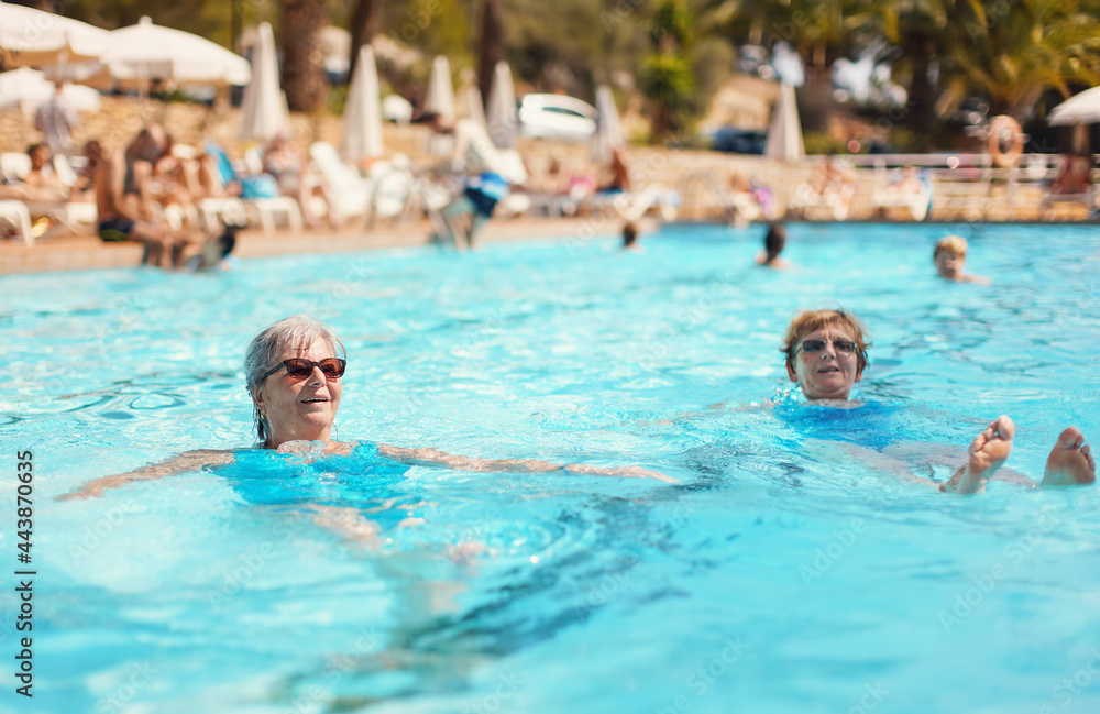 Two elderly senior women with grey hair, wearing blue swimsuit resting and swimming on their backs in hotel pool