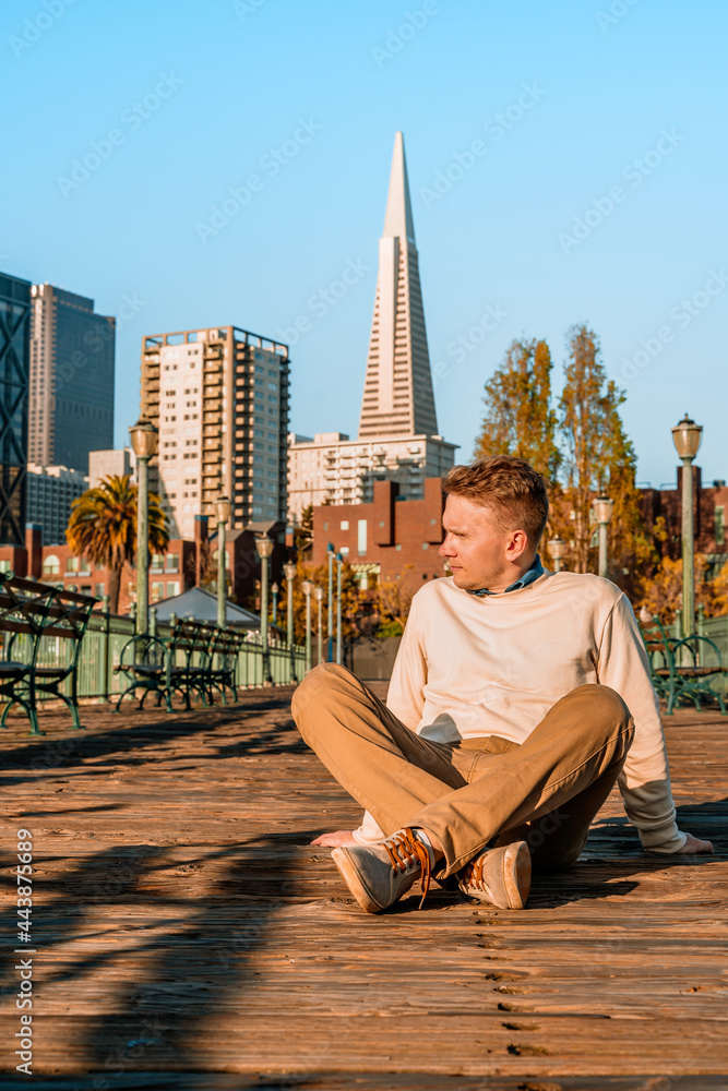 A young man sits on a wooden floor on a pier in the morning with a view of downtown and the Transamerica Tower in San Francisco