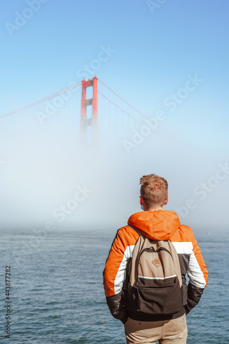 A young man with a jacket stands on the embankment and looks at the fog covering the Golden Gate Bridge in San Francisco, an incredible landscape © KseniaJoyg