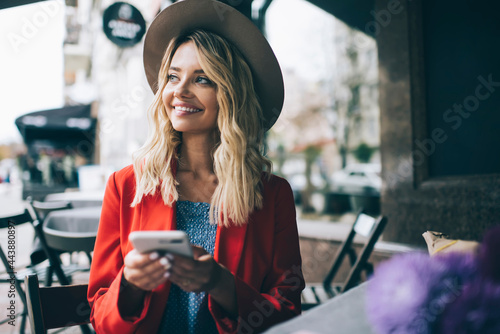 Cheerful female blogger with digital mobile technology using 4g internet during weekend time in street cafe, carefree hipster girl in trendy clothes holding smartphone device and smiling outdoors photo