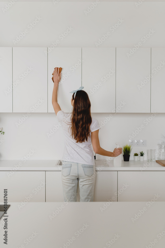 Rear view of young woman holding rag and spray bottle detergent cleaning kitchen. Rear view of young woman holding rag and spray bottle detergent cleaning kitchen.