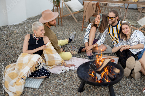 Young stylish friends sitting together by the fireplace outdoors. Talk and have fun during the picnic on the rooftop terrace