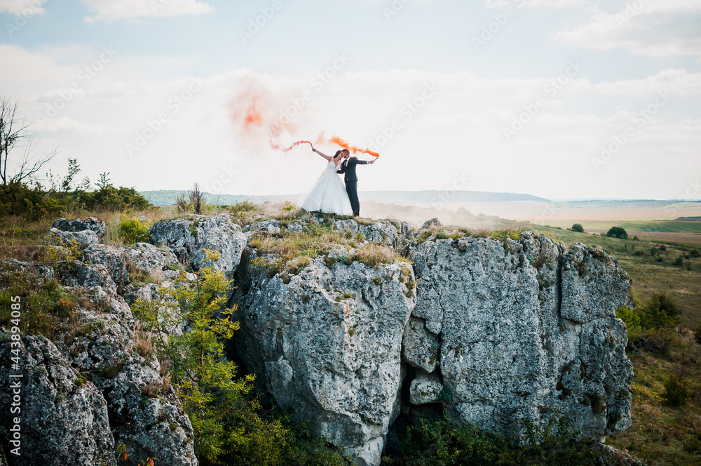 Newlyweds stand against the background of rocks and hold colorful smoke in their hands