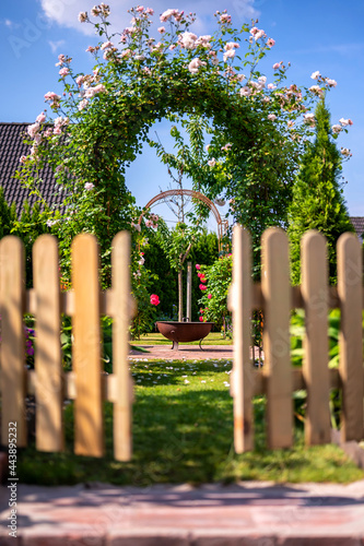 Fototapeta Naklejka Na Ścianę i Meble -  Offenes Gartentor mit Blick in einen wunderschönen Garten mit Rosenbogen und Feuerschale
