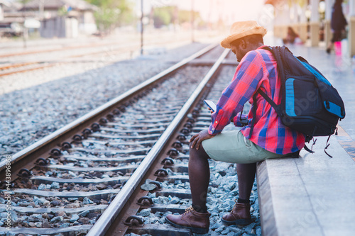 African male traveler waiting for the train on railway station with using smartphone. Adventure travel concept