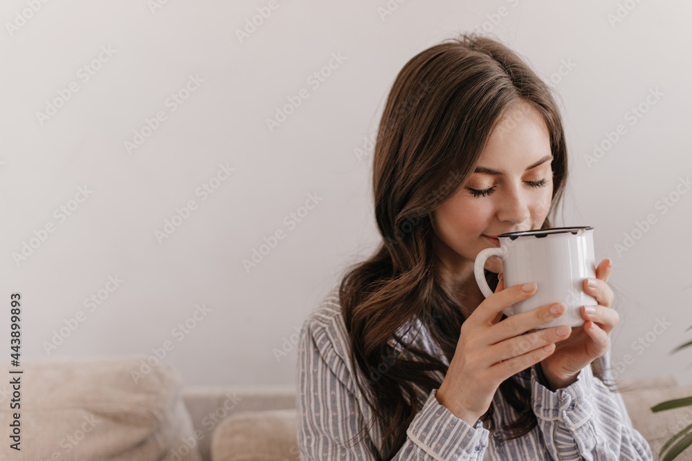Long-haired girl in blue pajamas enjoying aroma of tea. Woman holding big cup. Teen in shirt drinks coffee in morning at home sitting on sofa