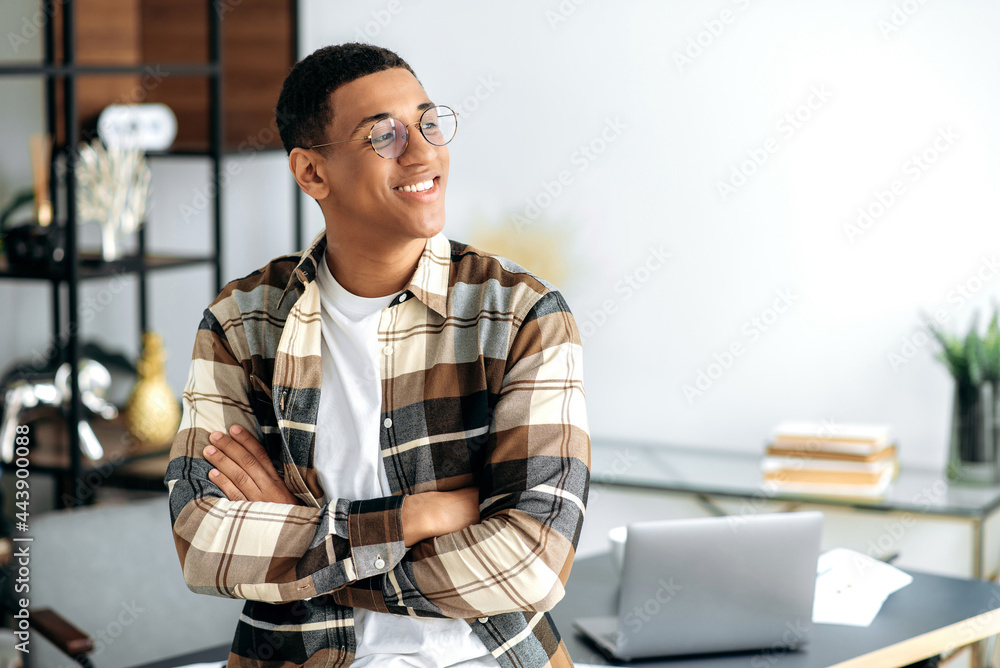 Fototapeta premium Portrait of an attractive confident positive young mixed race latino man with glasses, designer or freelancer, stylishly casual dressed, stand near a desk with arms crossed, looking to the side, smile