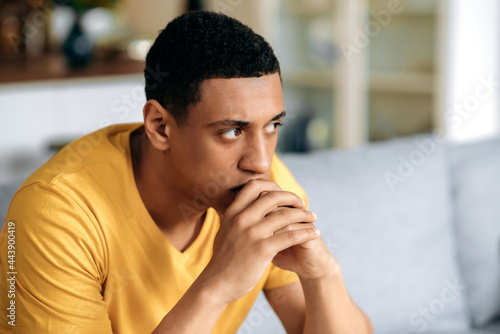 Frustrated unhappy latino millennial guy in a yellow t-shirt, sitting on the sofa in the living room, he has personal problems, failure at work, depression, bad mood, bad news, sad young man is lonely