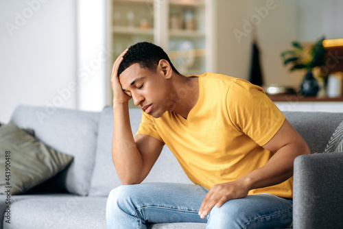 Frustrated sad handsome latino young alone man, sitting on a sofa at home, holding his forehead, having headache, depression, disappointed, closed eyes, needs psychological support and rest © Kateryna