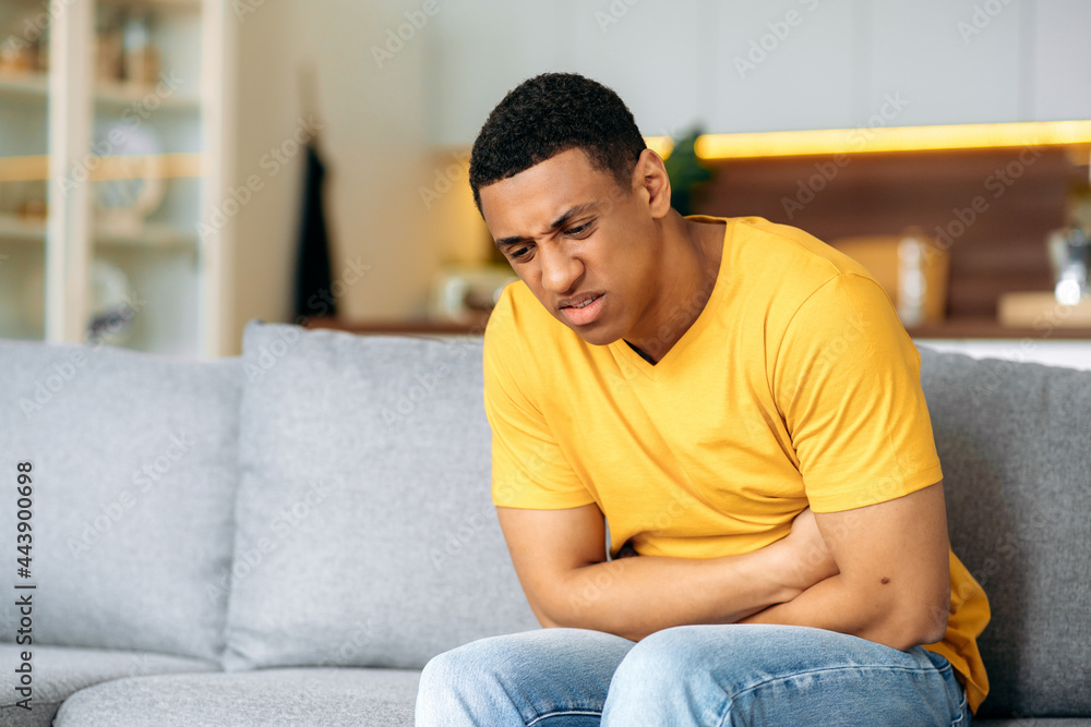 Hispanic frustrated unhealthy guy, sitting at home on the sofa, holding his stomach with his hands, suffering from stomach pain, stomach cramps, problems with the digestive tract, needs treatment Hispanic frustrated unhealthy guy, sitting at home on the sofa, holding his stomach with his hands, suffering from stomach pain, stomach cramps, problems with the digestive tract, needs treatment