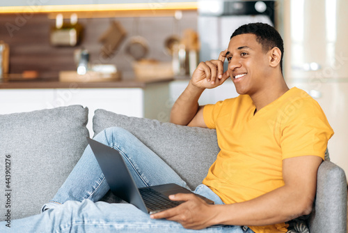 Positive latino guy sitting on a sofa at home in the living room, using a laptop, browsing the Internet, chatting online on social networks, makes online orders, looking to the side, smiling