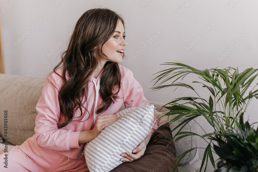 Charming girl with dark hair and gentle make-up looks to right on big flowerpot. Lady in pink sweatshirt holds striped pillow and sits on couch Charming girl with dark hair and gentle make-up looks to right on big flowerpot. Lady in pink sweatshirt holds striped pillow and sits on couch
