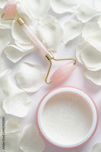 Face roller and jar of moisturizer on white background with rose petals. Facial skin care concept.
