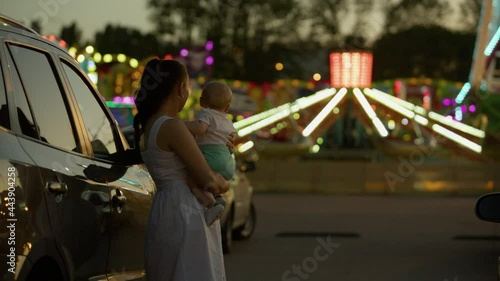 Mom and baby in a nighttime amusement park. Standing near a car holding an emotional child in her arms in a luna park where many lights in the background are out of focus.