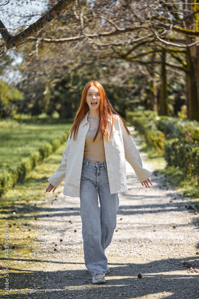 Positive caucasian female have fun in park, surrounded by green plants trees, smiling, looking at camera, in motion. Redhead lady in casual stylish clothes outfit, full-length portrait Positive caucasian female have fun in park, surrounded by green plants trees, smiling, looking at camera, in motion. Redhead lady in casual stylish clothes outfit, full-length portrait