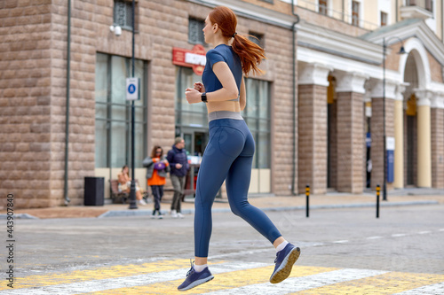 female Runner athlete running at road. woman fitness jogging workout wellness concept. beautiful lady with natural red hair in blue sportive outfit engaged in sport, wprkout in city