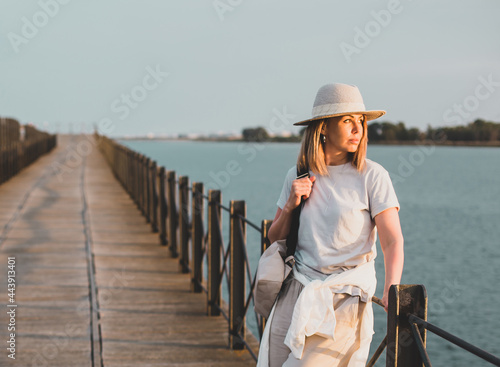 A girl in light clothes and a hat is standsing on the pier and looking at the sunset against the backdrop of the seascape. Huelva, Spain.