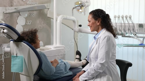 Cute preadolescent mixed race girl and woman stomatologist communicating in denal office before examination. Friendly pediatric dentist giving high five little patient sitting on stomatological chair