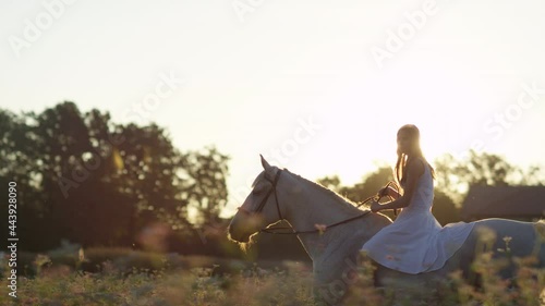 SLOW MOTION, LENS FLARE: Carefree girl wearing a white sundress rides her beautiful mare bareback on a sunny summer evening. Young Caucasian woman horseback riding in the golden lit countryside. photo