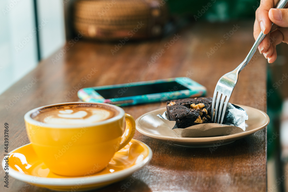 Hands of a young woman with a fork . A plate of cake and smartphone on the table. In the foreground is a cup of coffee