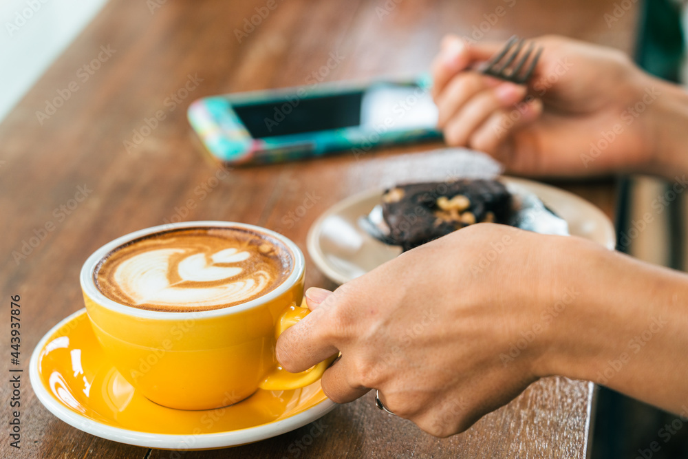 Young woman's hand with a cup of coffee. In the background, a smartphone on the  wooden table and a cake in a plate in cafe