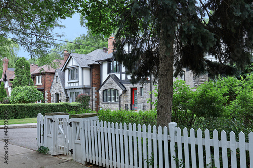 Tree lined residential street with older two story Tudor style houses and white picket fence around front yard