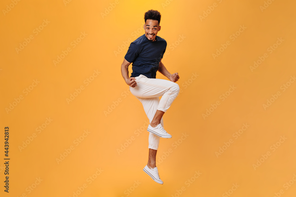 Stylish guy in white pants and dark T-shirt jumping on isolated background. Full-lenght portrait of man moving on orange backdrop Stylish guy in white pants and dark T-shirt jumping on isolated background. Full-lenght portrait of man moving on orange backdrop
