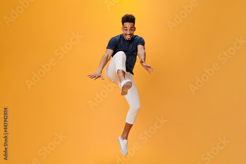 Stylish guy emotionally poses and jumps on orange background. Snapshot of handsome dark-haired man in white pants moving on isolated