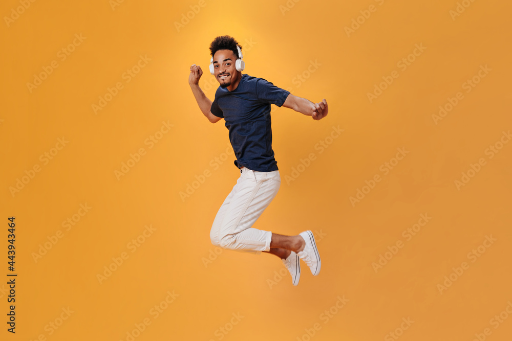 Energetic man in stylish outfit jumping on orange background and listening to music on headphones. Cool guy un blue shirt having fun on isolated backdrop
