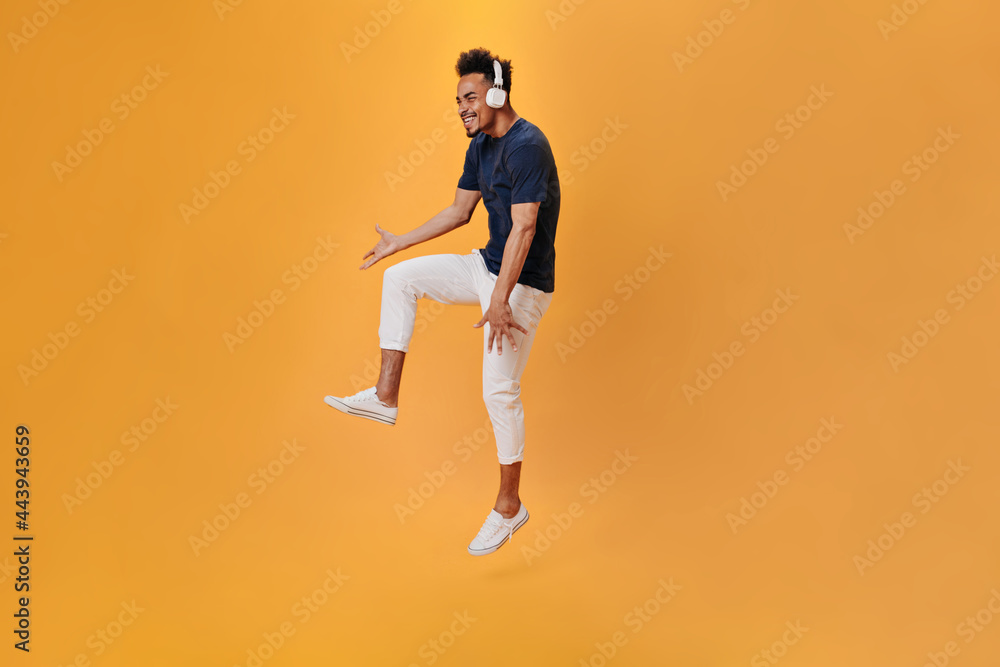 Guy in white pants listens to music with headphones and jumps on orange background. Cheerful man in dark blue shirt dancing on isolated backdrop