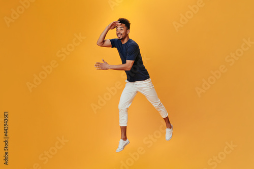 Dark skinned guy in black and white outfit jumping on isolated background. Brunette man in tee moving on orange backdrop © Look!