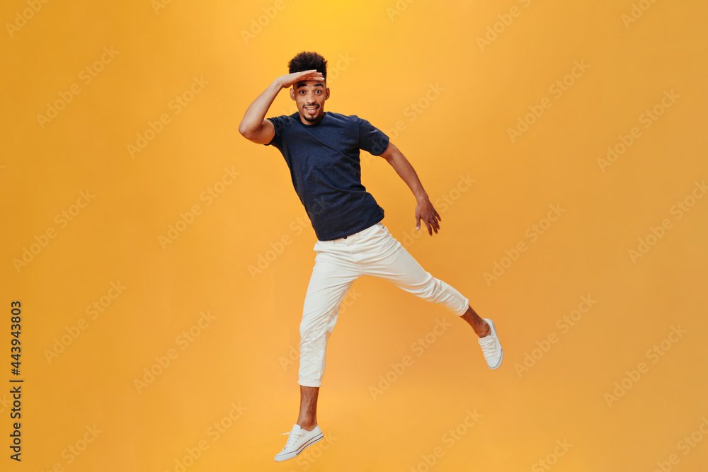 Cheerful guy jumps on orange background and puts his palm to his forehead. Full-lenght man in white pants and tee posing on orange background