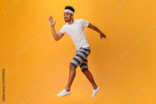 Dark-skinned guy in shorts and T-shirt runs on orange background. Sportsman has workout on isolated. Portrait of dark-skinned teen