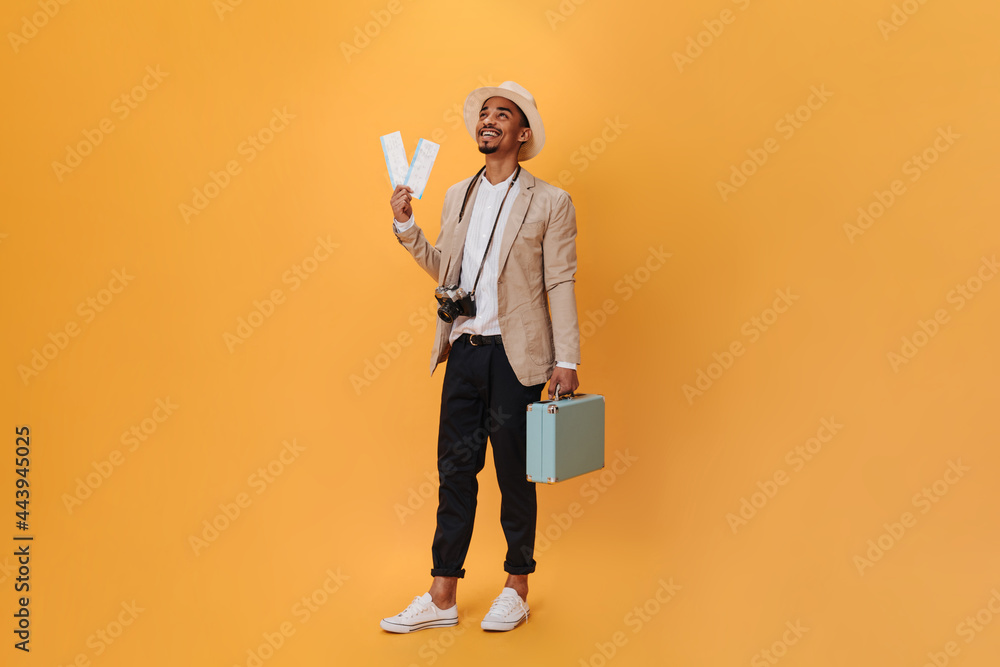 Handsome young guy posing in studio on isolated. Charming happy man in black pants and beige jacket posing with suitcase and tickets Handsome young guy posing in studio on isolated. Charming happy man in black pants and beige jacket posing with suitcase and tickets