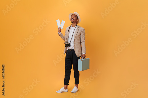 Handsome young guy posing in studio on isolated. Charming happy man in black pants and beige jacket posing with suitcase and tickets