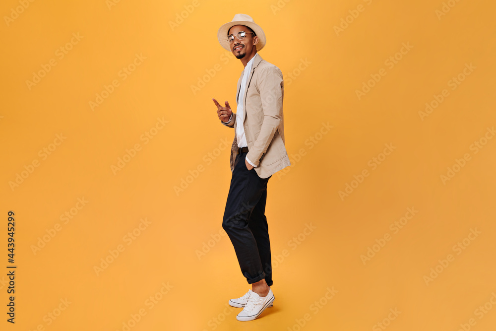 Guy in hat and suit posing on isolated background. Portrait of dark-skinned man in black pants, white tee and beige jacket on orange backdrop Guy in hat and suit posing on isolated background. Portrait of dark-skinned man in black pants, white tee and beige jacket on orange backdrop