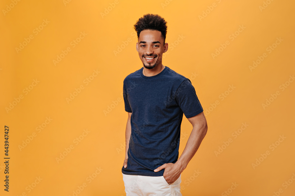 © Look! - Man in black t-shirt smiles sweetly on orange background. Portrait of guy in blue tee posing on isolated backdrop