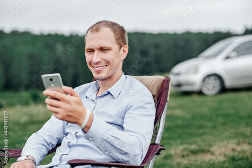 young man freelancer sitting on chair and using smart phone at camping site in forest or meadow. Remote work and outdoor activity in summer.