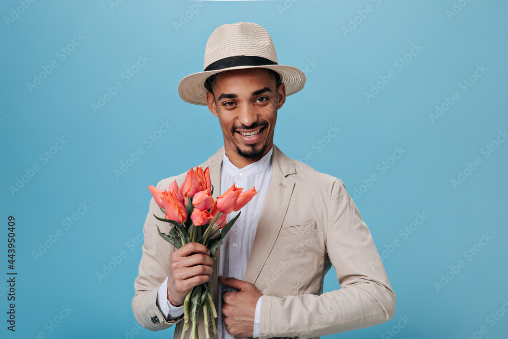 Stylish man in suit and hat holding bouquet on blue background. Porprait of guy in beige outfit smiling and posing with tulips on isolated backdrop Stylish man in suit and hat holding bouquet on blue background. Porprait of guy in beige outfit smiling and posing with tulips on isolated backdrop