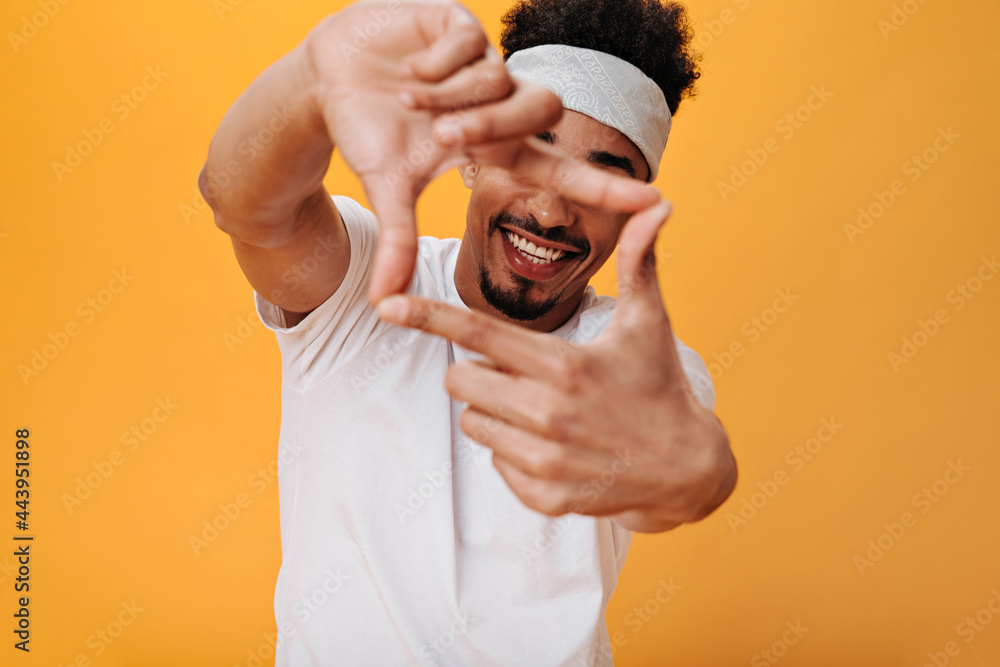 Guy puts his fingers in shape of rectangle. Man in white tee and headband smiling and posing happily on orange isolated background Guy puts his fingers in shape of rectangle. Man in white tee and headband smiling and posing happily on orange isolated background