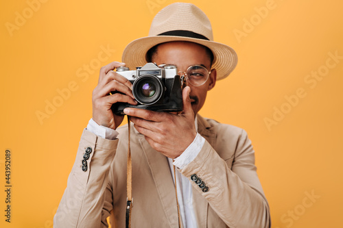 Man in stylish hat and beige suit holding retro camera. Portrait of charming guy in eyeglasses and white tee taking photo on orange background