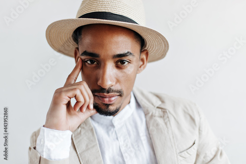 Guy in white shirt and beige jacket looks into camera on white background. Close-up portrait of man in suit and hat posing on isolated backdrop