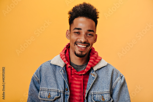 African American man in denim jacket smiling on orange background. Joyful guy in red hoodie posing hapily on isolate backdrop photo