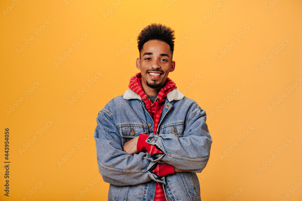 Young guy in denim jacket is smiling on orange background. Man in red hoodie  looking into camera. Portraitof positive teen on isolated Stock Photo