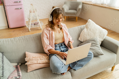 Image of young pleased happy cute beautiful  woman sit indoors on sofa  using laptop computer listening music with earphones  in living room.