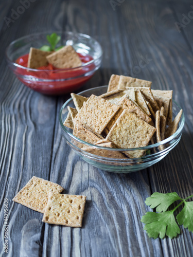 Natural crunchy mini square-shaped bread in a glass bowl on a dark wooden background. Dipping crispbreads in ketchup in the background. Wood texture. Diet snack