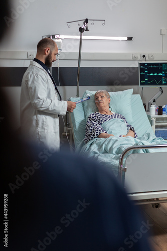 Doctor questioning senior woman laying in hospital bed  breathing with help from oxygen tube because of lungs surgery. Patient recieving medicine through intrevenous line.